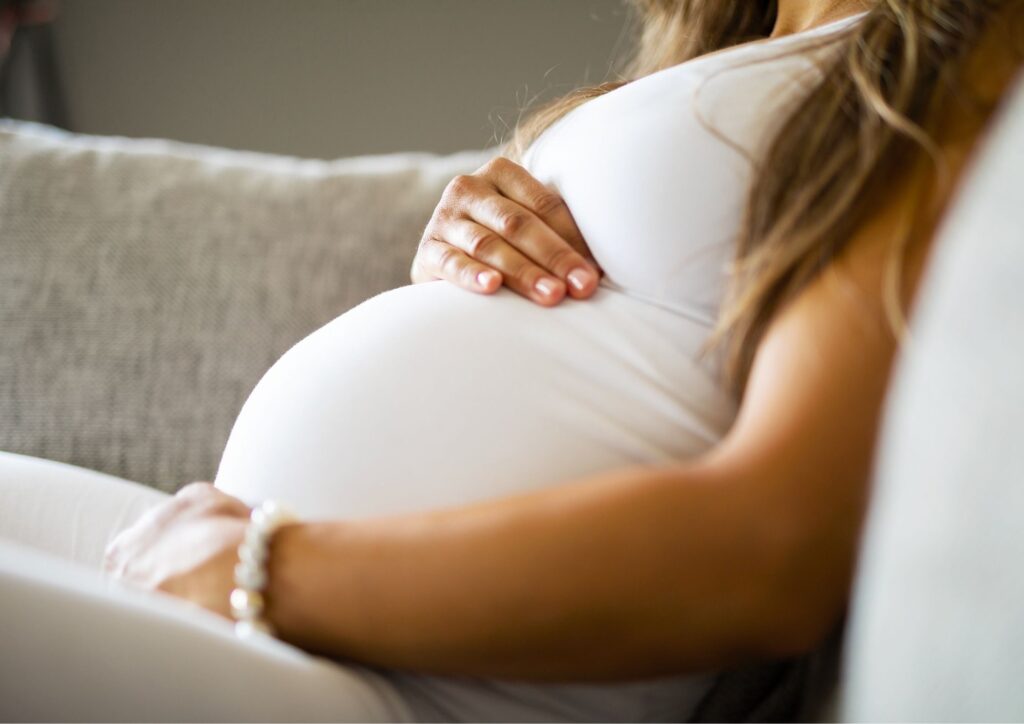Pregnant woman sitting on a couch in warm, soft light, gently holding her belly during a quiet, peaceful moment.
