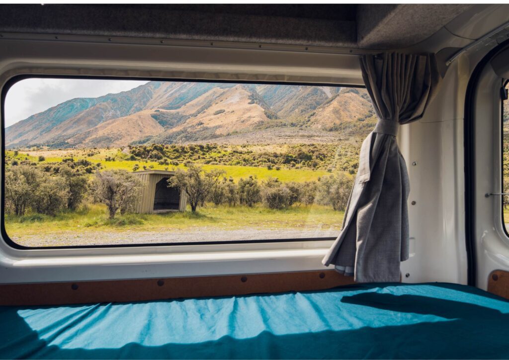 A mountain landscape seen through the back of a campervan with a bed, capturing the freedom and simplicity of solo travel in New Zealand.