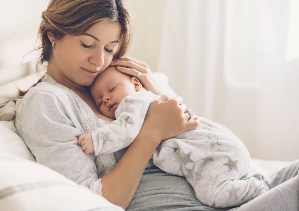 A mother half‑sitting in bed, holding her sleeping baby on her chest. Both have their eyes closed, creating a soft, intimate moment.