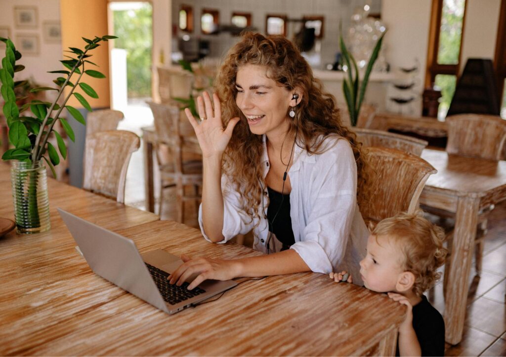 A mother sitting at a kitchen table, smiling and waving at someone on her laptop during a video call, while her toddler’s face pops into the frame beside her.