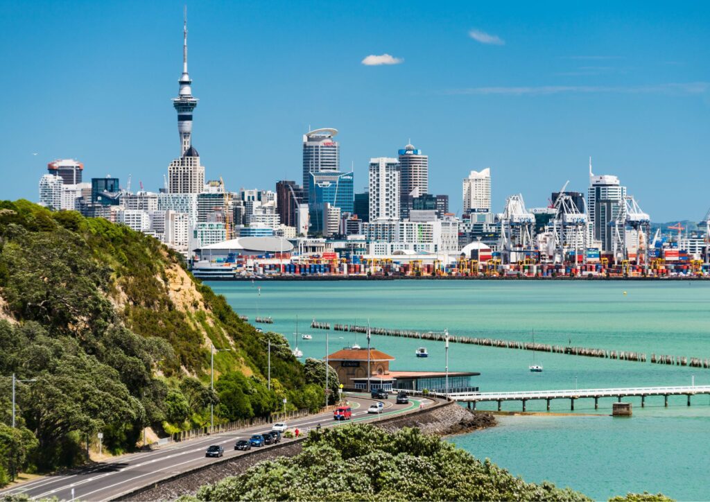 A wide view of Auckland from the Orākei direction, showing the city skyline with the Sky Tower rising above the buildings and the blue ocean in the foreground.