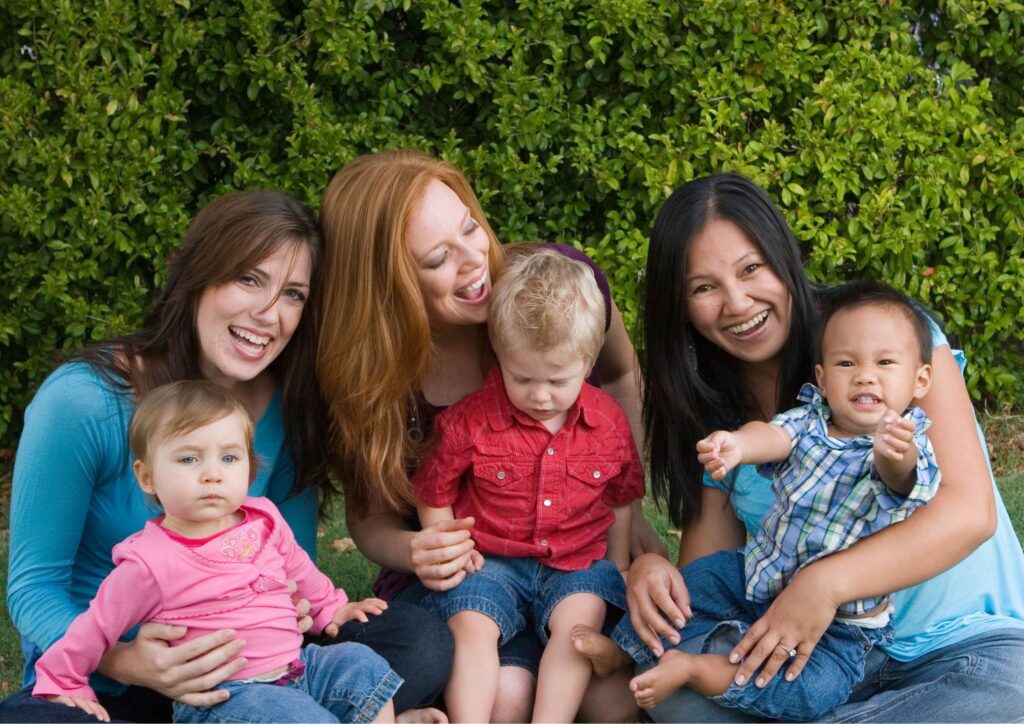 Three mums sitting together with their toddlers during an outdoor playdate, talking while the children play nearby.