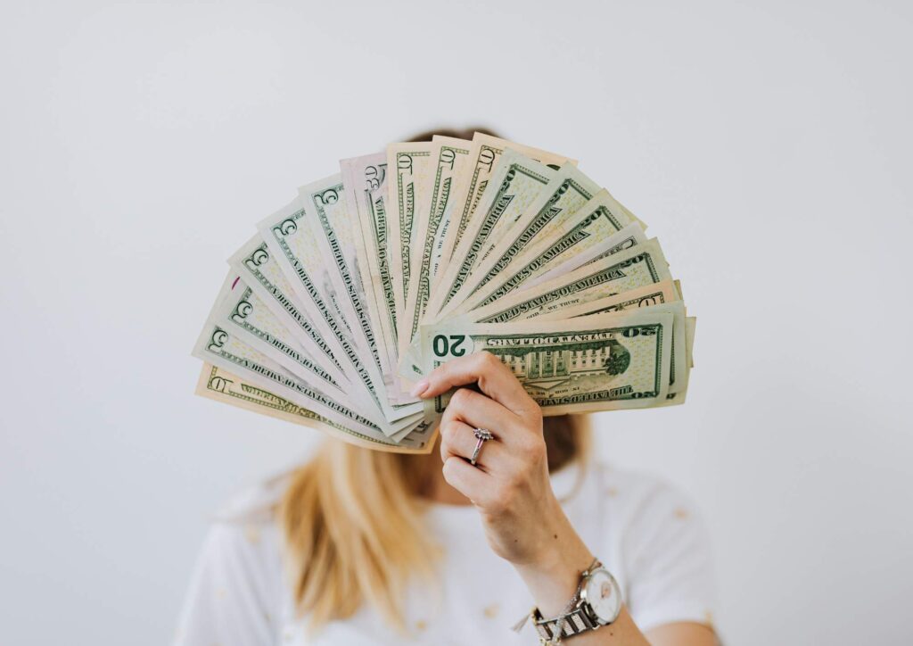 A person holding a fan of cash in front of their face, symbolizing the pressure to find side hustles and the rise of platforms like OnlyFans as financial options.