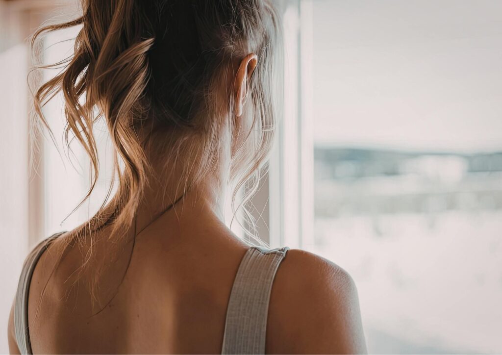 A woman standing by a window, looking outside in soft light, reflecting the emotional experience of homesickness and rebuilding identity abroad.