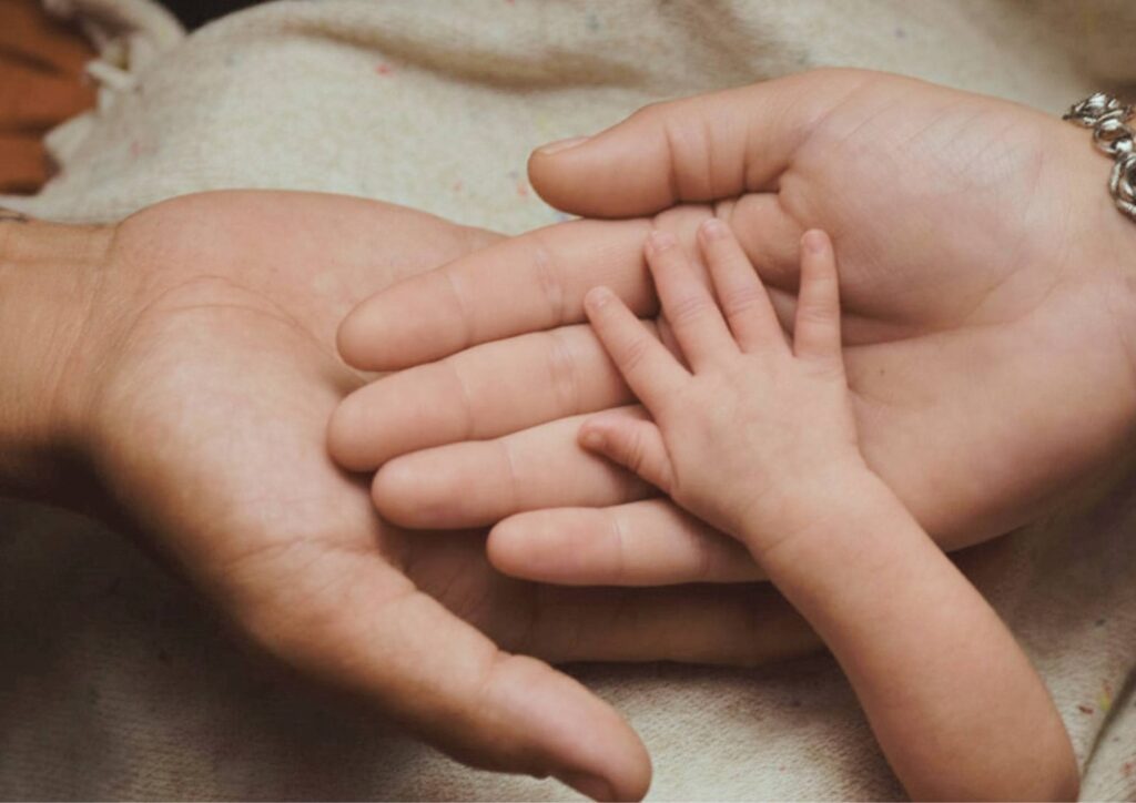 A close-up of three hands layered together — an adult’s hand, a smaller hand, and a baby’s hand — symbolizing protection, connection, and the responsibility of caring for a child.