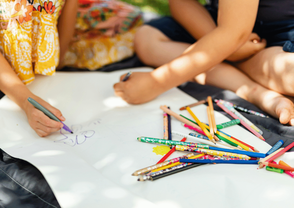 Children sitting on the ground colouring with bright pencils on a large sheet of paper.