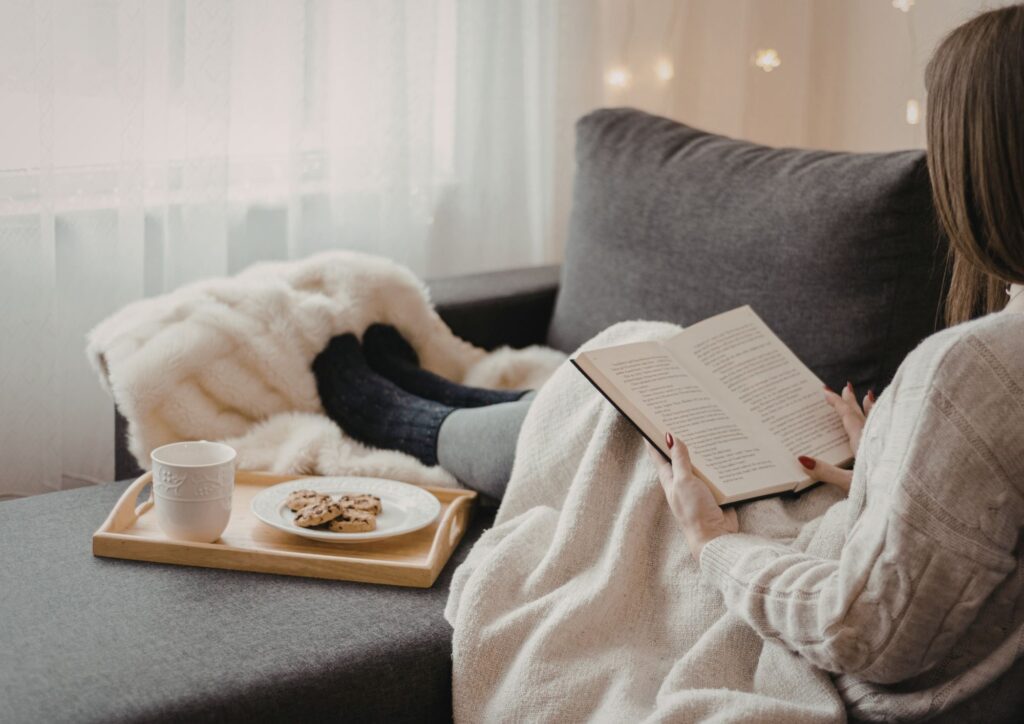 A mother sits curled up on a grey sofa under a soft blanket, holding an open book. A tray with a warm drink and cookies rests beside her, creating a cozy scene that reflects the longing for rest and small moments of self‑care.