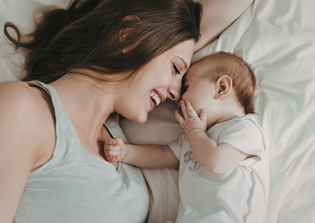 A mother lies on a bed facing her baby, their noses touching as the baby rests a hand near her face. The moment captures tenderness, connection, and the deep love that makes the challenges of motherhood worthwhile.