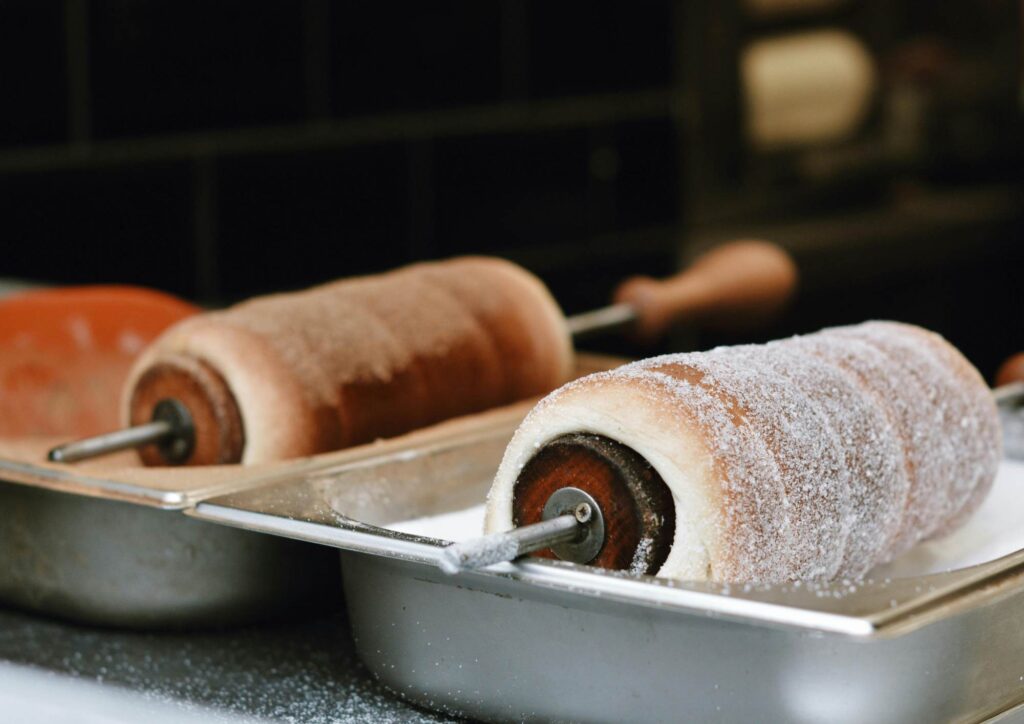 Two freshly baked chimney cakes cooling on wooden rollers, coated in caramelised sugar inside a metal tray.