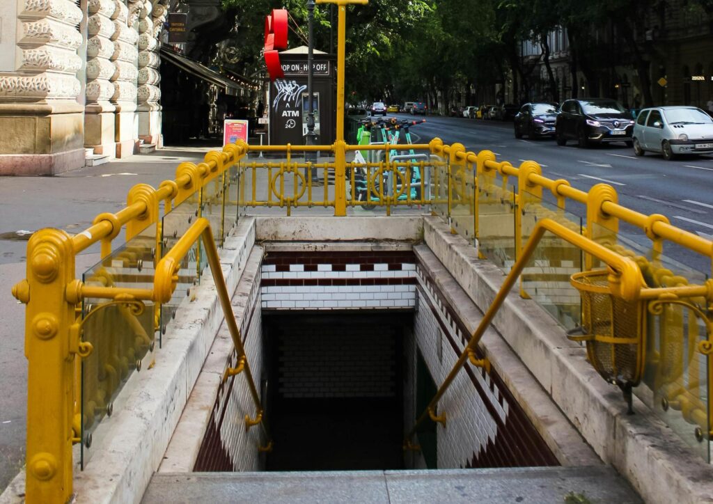 A Budapest metro entrance with bright yellow railings and tiled walls, leading down into the underground station on a busy city street.