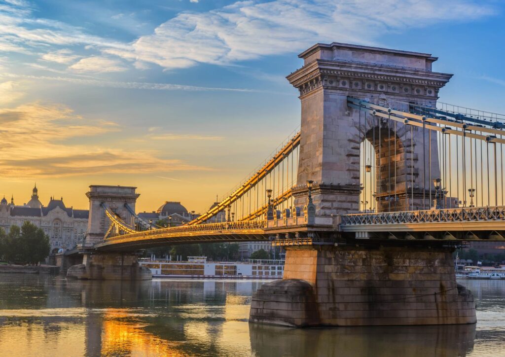 The Széchenyi Chain Bridge glowing in warm sunset light, spanning the Danube with historic buildings lining the riverbank.