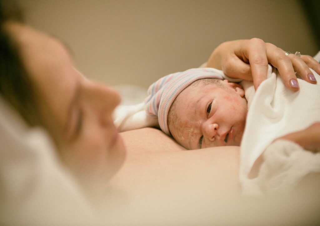 Newborn lying on an adult’s chest immediately after birth, wrapped in a blanket and wearing a striped cap, with the adult’s hand gently touching the baby.