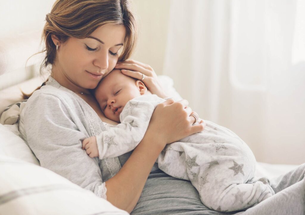 Adult holding a sleeping infant close to their chest on a bed or couch, both resting peacefully in soft daylight.