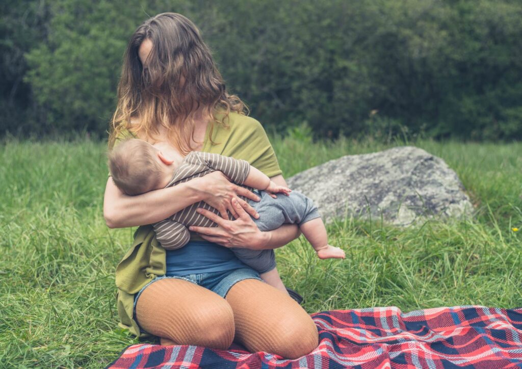 Adult breastfeeding an infant outdoors on a plaid blanket, surrounded by greenery and soft natural light.