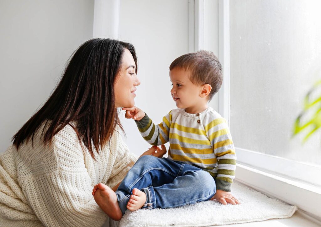 Adult sitting with a toddler by a bright window, the toddler gently touching the adult’s chin in a warm, intimate moment.