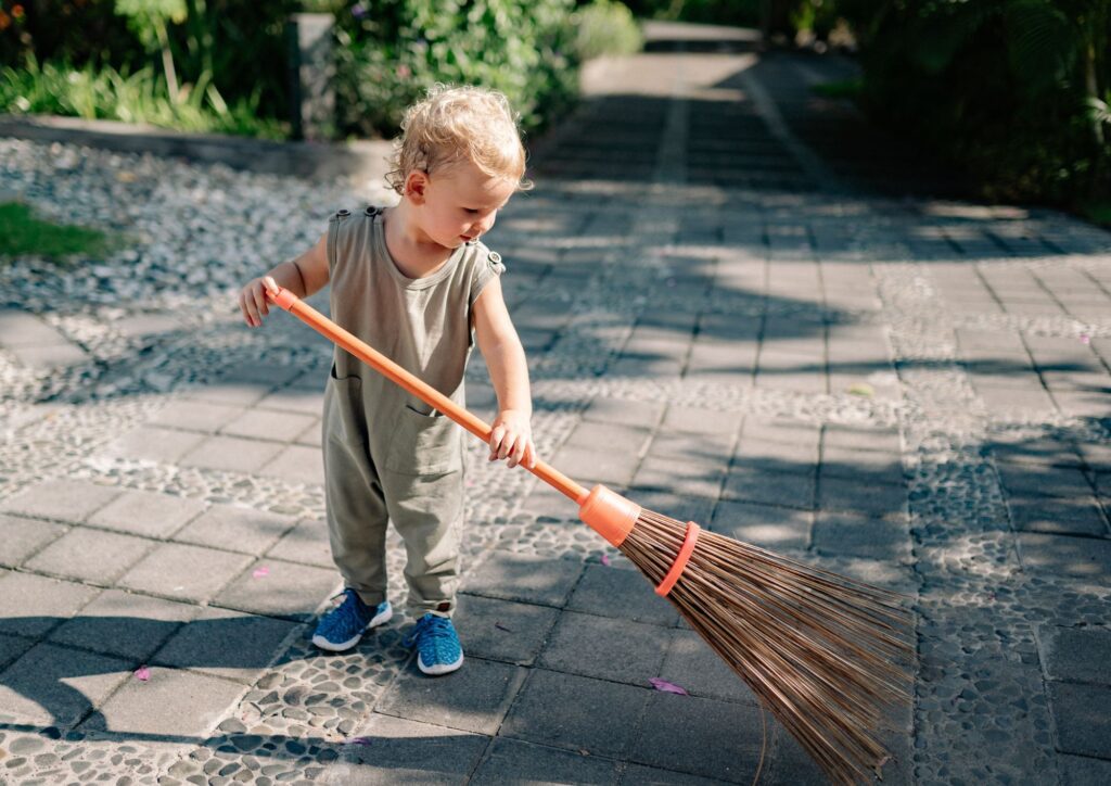 A toddler sweeping an outdoor walkway with a small broom, learning through imitation and everyday household rituals.