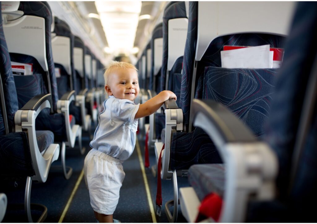 A toddler standing in the airplane aisle holding onto a seat armrest.