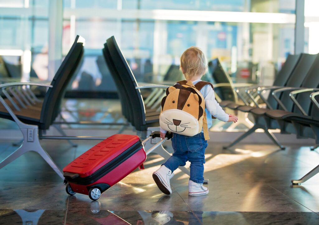 A toddler walking through an airport pulling a small suitcase and wearing a lion backpack.