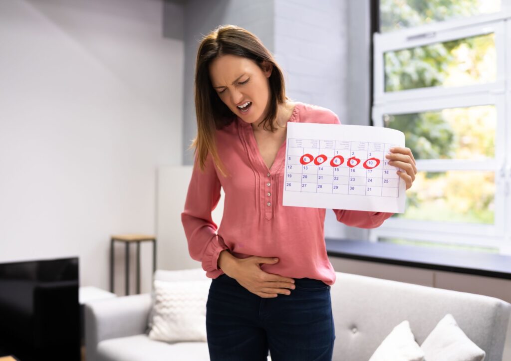 A woman holding a calendar with several dates circled in red while touching her lower abdomen, showing awareness of her menstrual cycle and its impact on daily life.