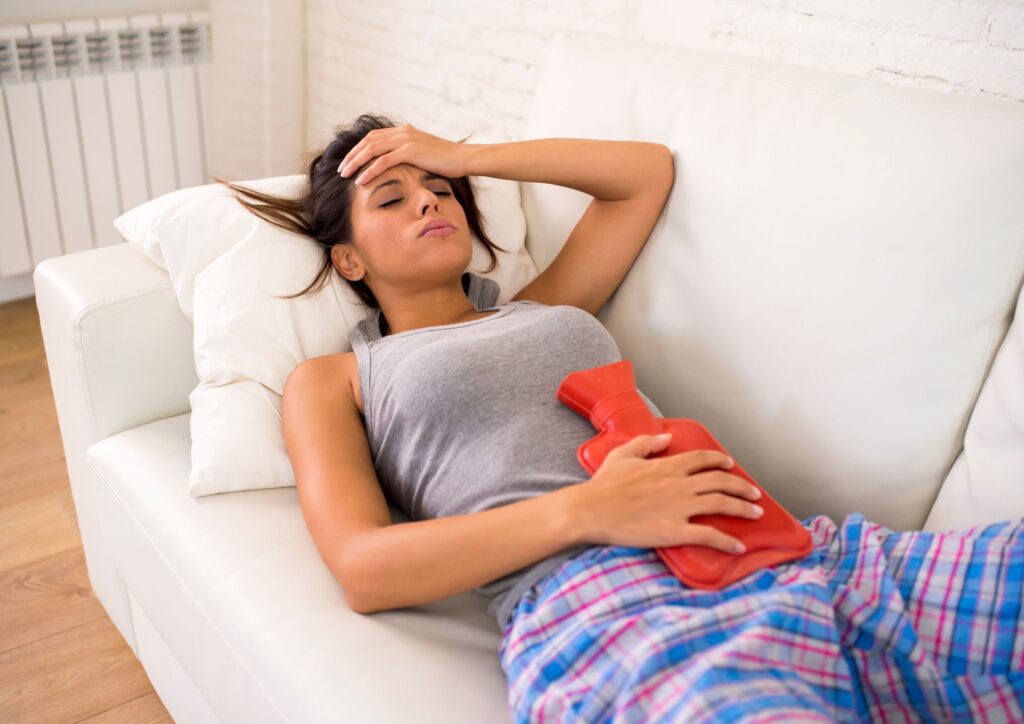 A woman lying on a couch holding a red hot water bottle against her lower abdomen, illustrating period cramps and self-care.