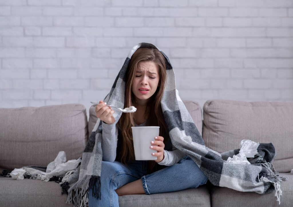 A woman sitting on a couch with a blanket over her head, holding a spoon and tissues around her, showing exhaustion and seeking comfort during her period.