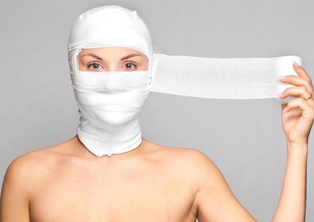 A person with their head wrapped in white gauze bandages, representing extreme cosmetic procedures or transformation.
