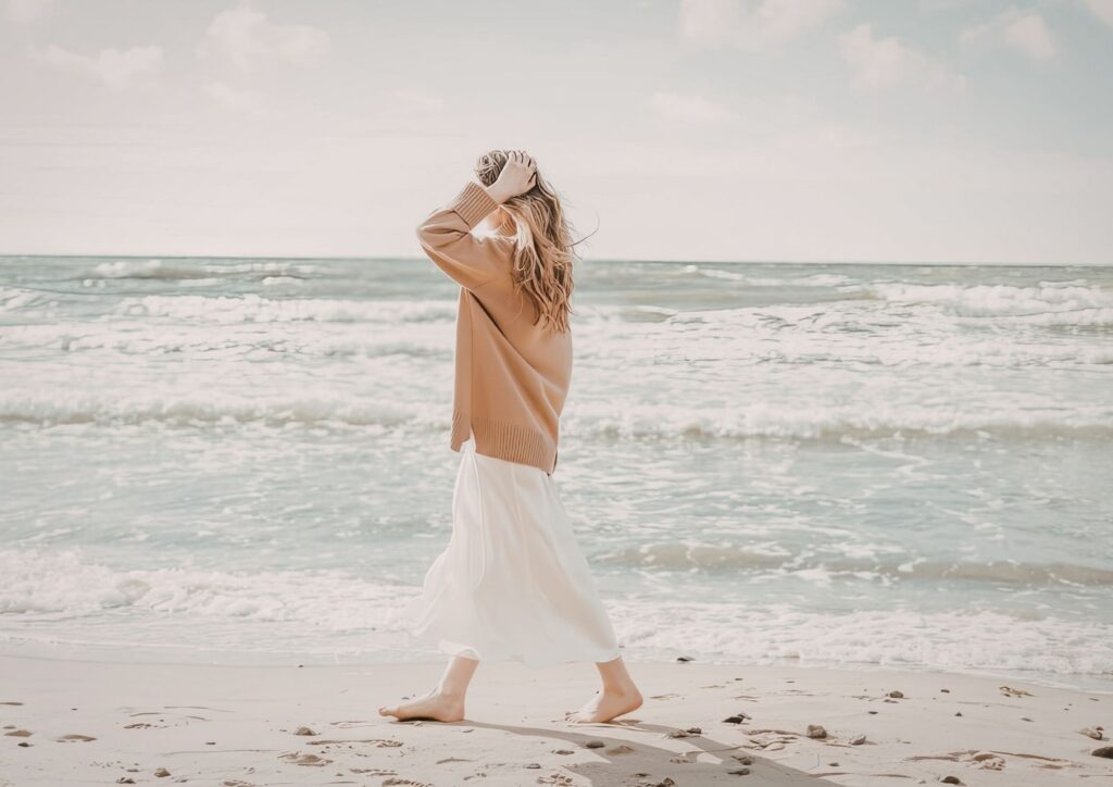 Person walking barefoot along a sandy beach in warm weather, wearing a light sweater and skirt, with gentle ocean waves in the background.