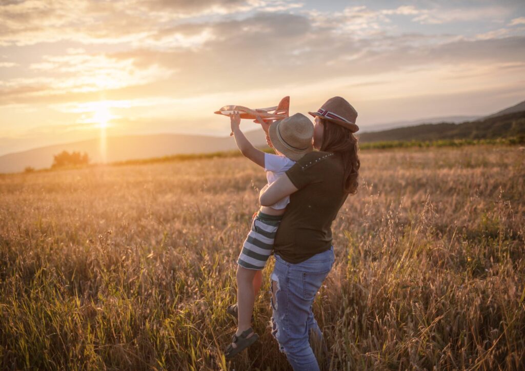 A parent holding a child in a grassy field at sunset while the child lifts a toy airplane toward the sky.