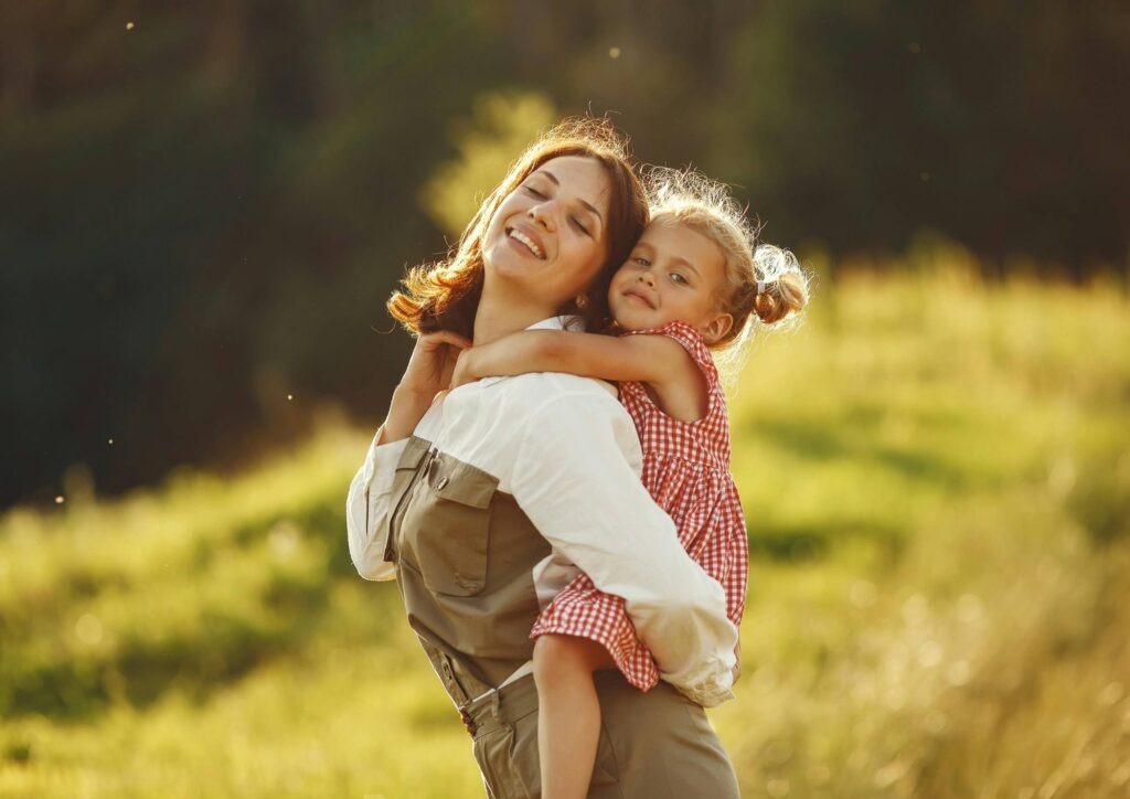 A mother holding her daughter in a sunlit field, both wrapped in warm golden light.