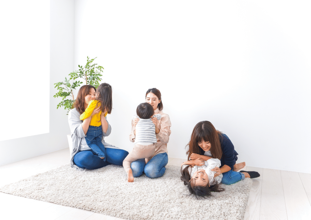 Three adults sitting on a carpeted floor, each playing closely with a toddler in a bright, minimal room.