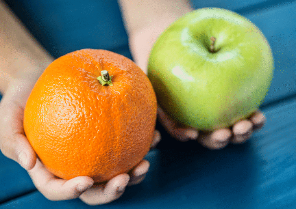 Two hands holding an orange and a green apple against a blue background, symbolizing comparison between different things.