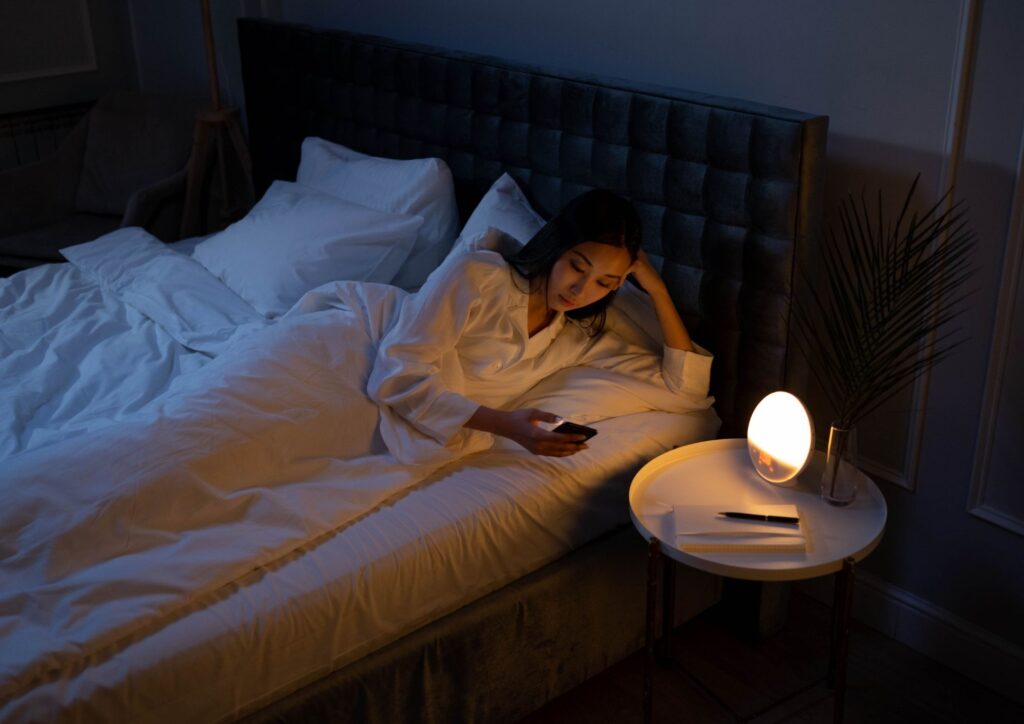 Person lying in bed looking at a mobile phone in a dimly lit room.